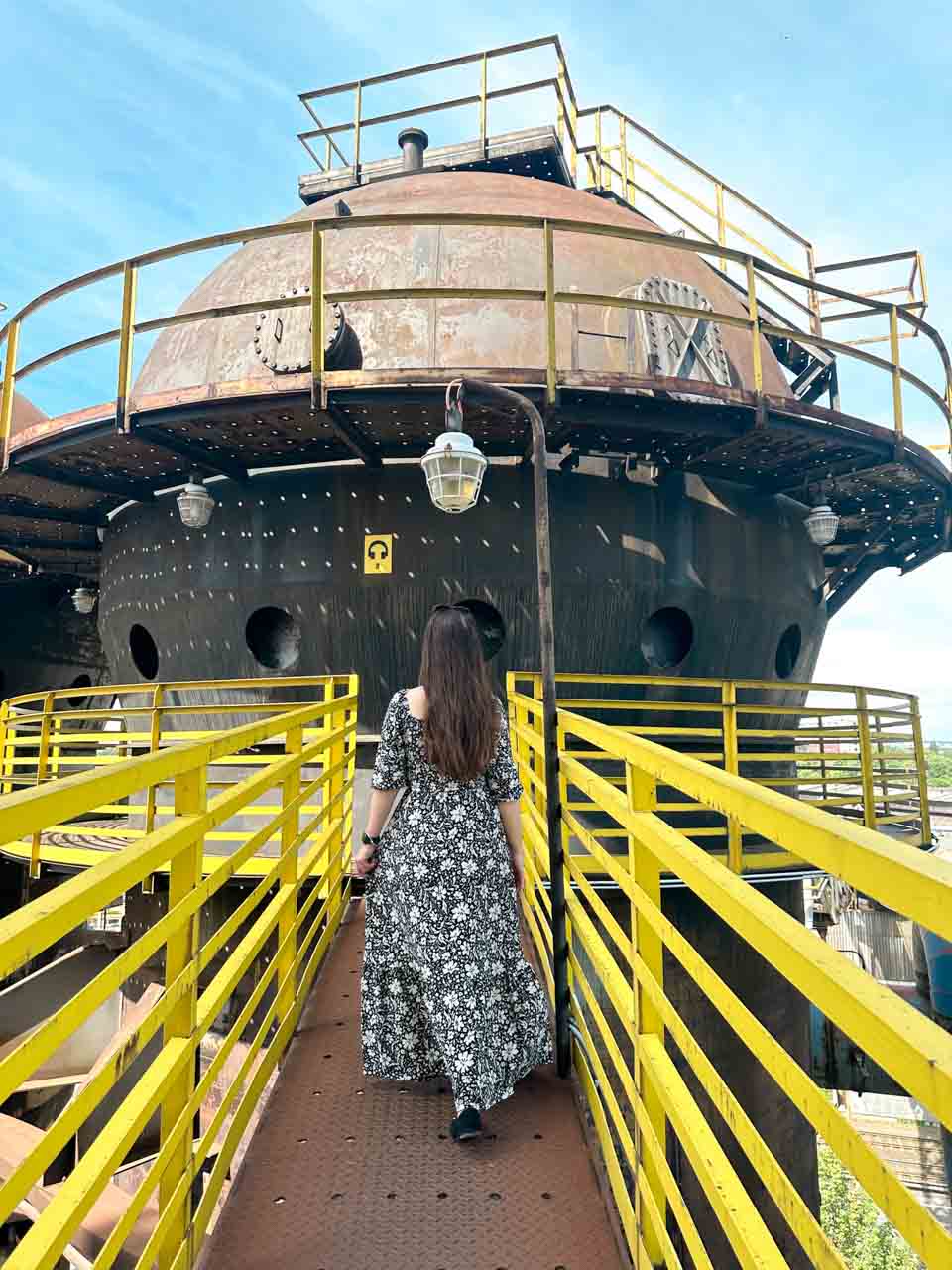 Woman in a black and white maxi dress walking towards industrial tanks on a yellow walkway at the Dolní Vítkovice complex in Ostrava