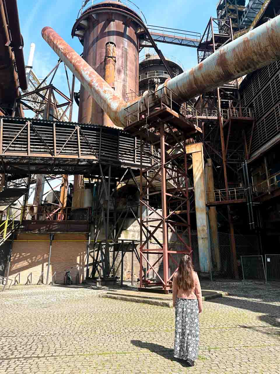 Woman in a black and white maxi dress and a pink jacket facing an imposing rusty industrial structure inside the Dolní Vítkovice complex in Ostrava