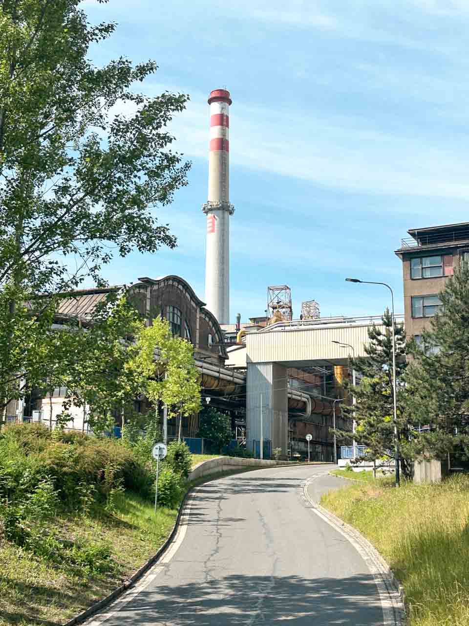 Tree-lined road leading to an industrial facility with a tall chimney in Ostrava