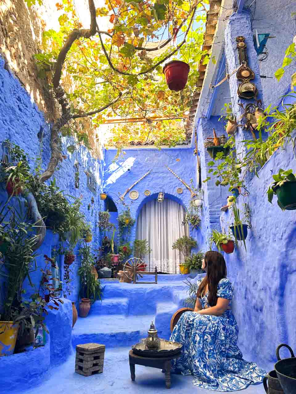 A woman in a blue floral dress sitting in a serene blue courtyard with hanging plants in Chefchaouen, Morocco