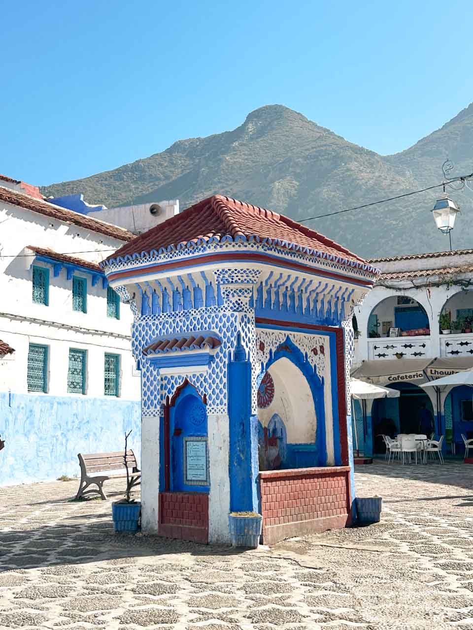 A vibrant white, blue, and red water fountain in El Haouta Square in Chefchaouen, with mountain peaks rising in the background