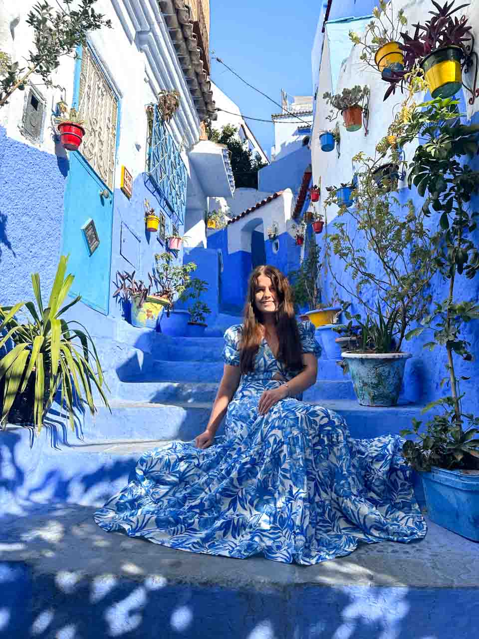 A woman in a blue floral dress sitting on vibrant blue steps, surrounded by colourful potted plants