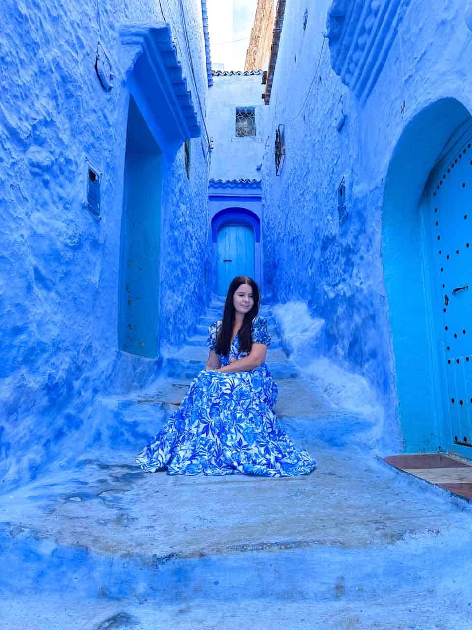 A woman in a blue floral dress seated on blue steps, surrounded by vibrant blue walls and doorways in Chefchaouen, Morocco