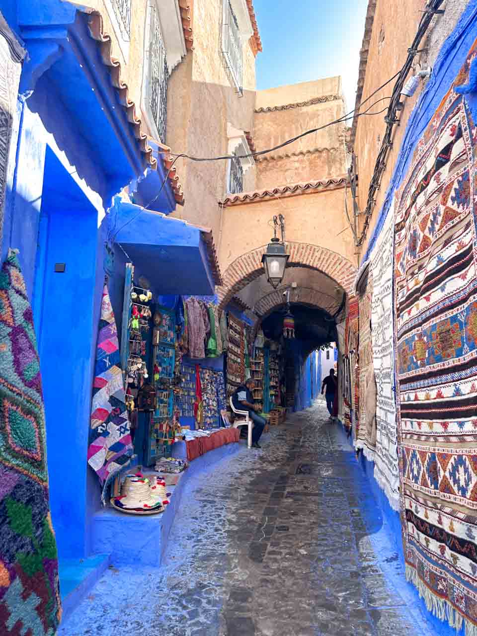 A narrow alley lined with colourful textiles, rugs, and crafts displayed outside shops under a brick archway in Chefchaouen, Morocco