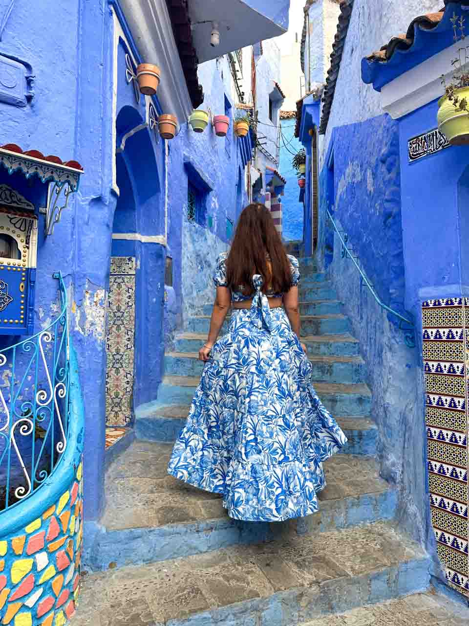 A woman in a blue floral dress walking up colourful steps in a blue alley lined with hanging plants
