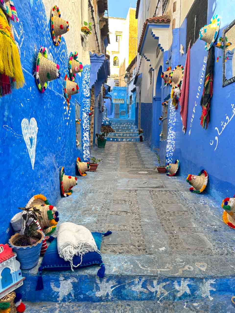 A colourful alley adorned with traditional woven hats and textiles, leading up blue-painted steps