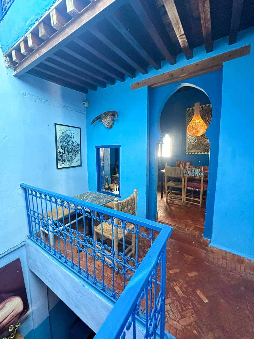 A riad interior with blue walls and wooden beams, featuring a central courtyard overlooking a room below, with rustic chairs and tables
