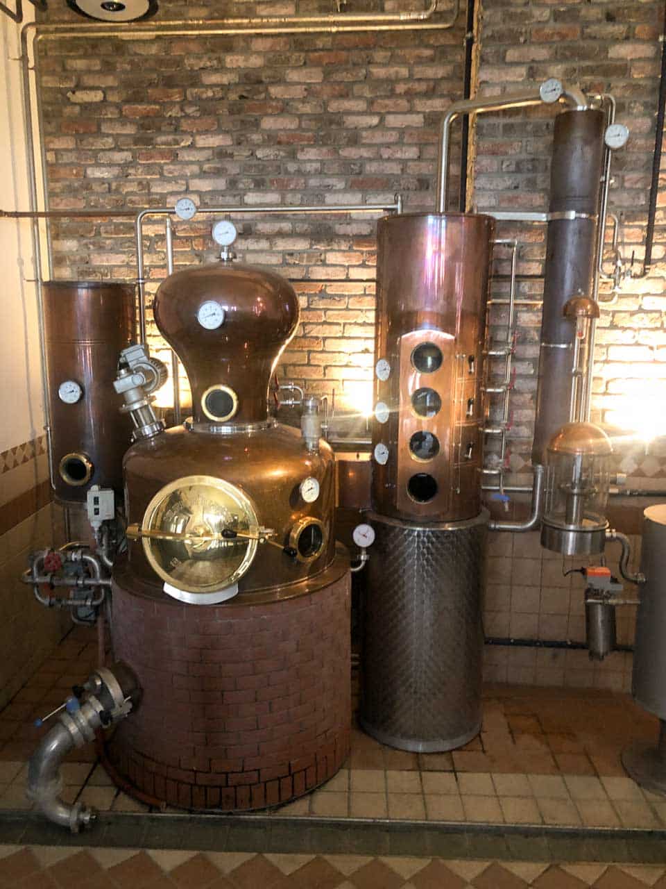 A copper distillation apparatus with gauges and pipes set against a rustic brick wall inside the Hausbrauerei Altstadthof brewery in Nuremberg, Germany