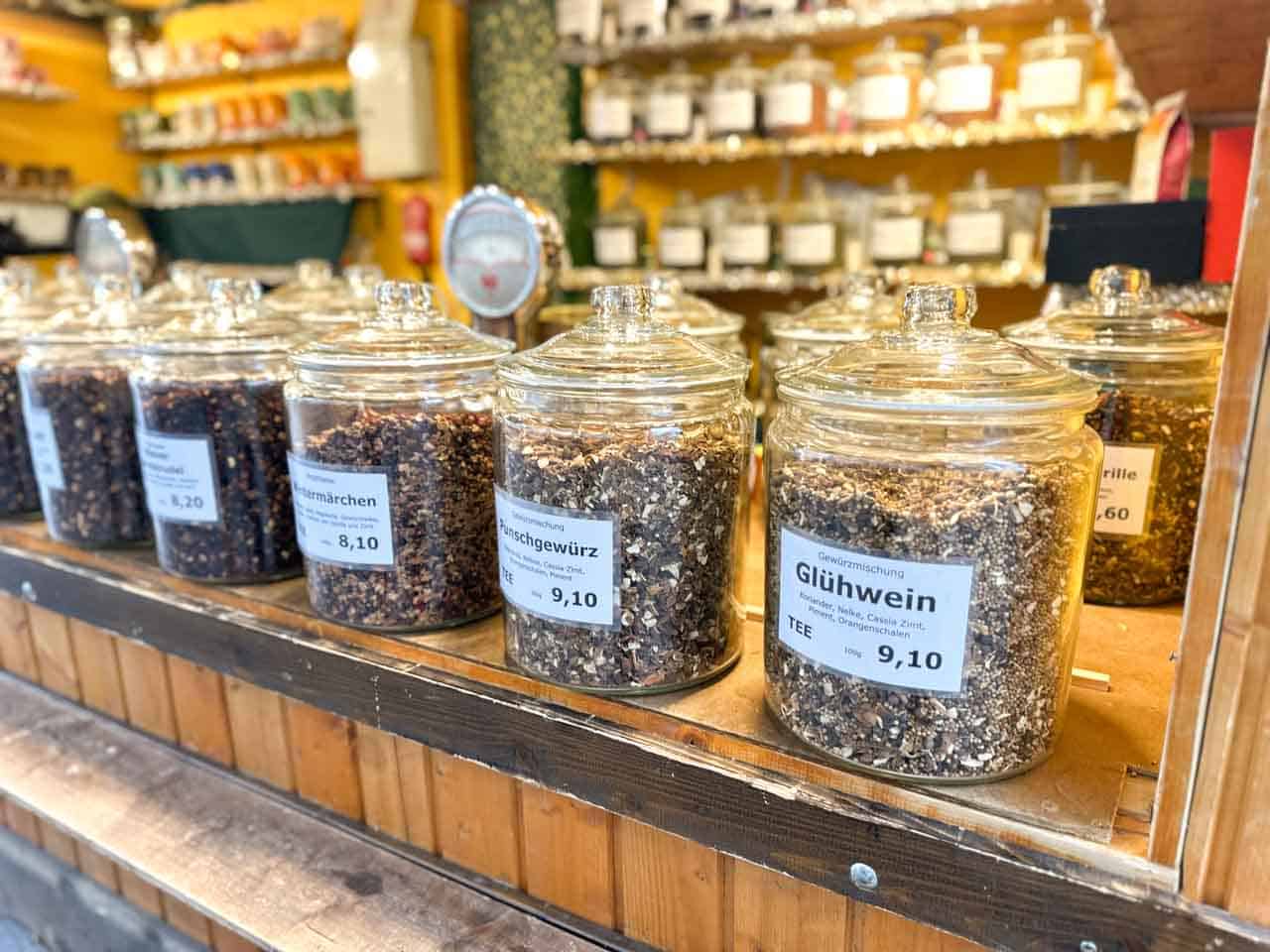 Glass jars filled with loose tea blends like Glühwein and Punch spices, lined up on a rustic wooden Christmas market counter in Vienna