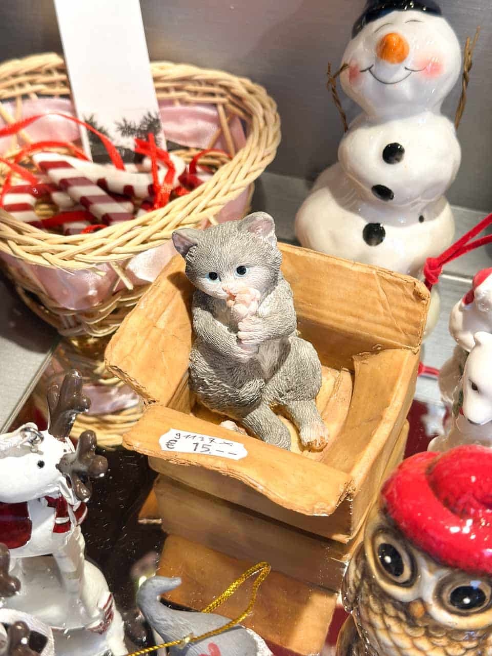 A small grey kitten figurine sitting in a wooden box, surrounded by festive decorations and a snowman ornament