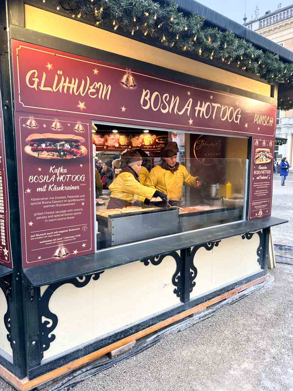 A Schönbrunn Christmas market stall selling Glühwein and Bosna hot dogs, with staff in yellow coats preparing food