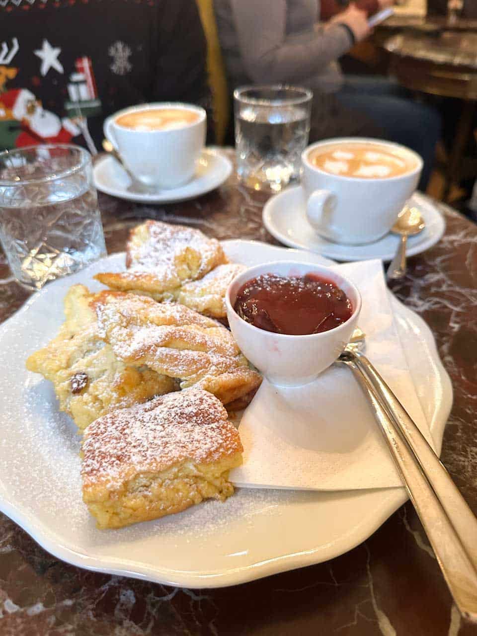 A plate of Kaiserschmarrn topped with powdered sugar, served with jam, alongside two cups of coffee on a marble table at Café Demel in Vienna