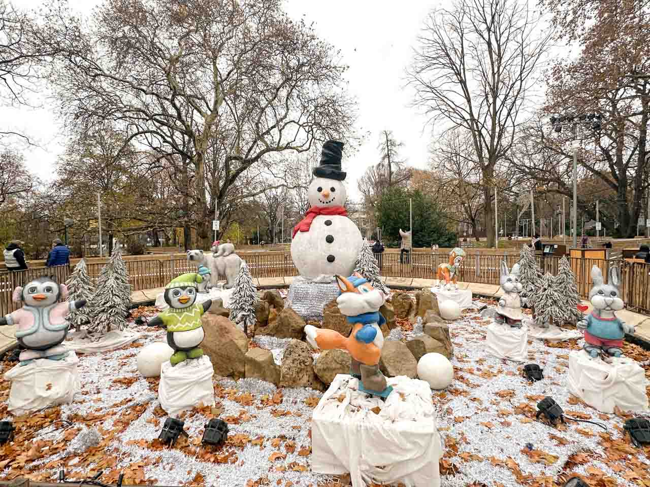 A winter display with a snowman and animal statues surrounded by frosted trees and festive decorations in a park