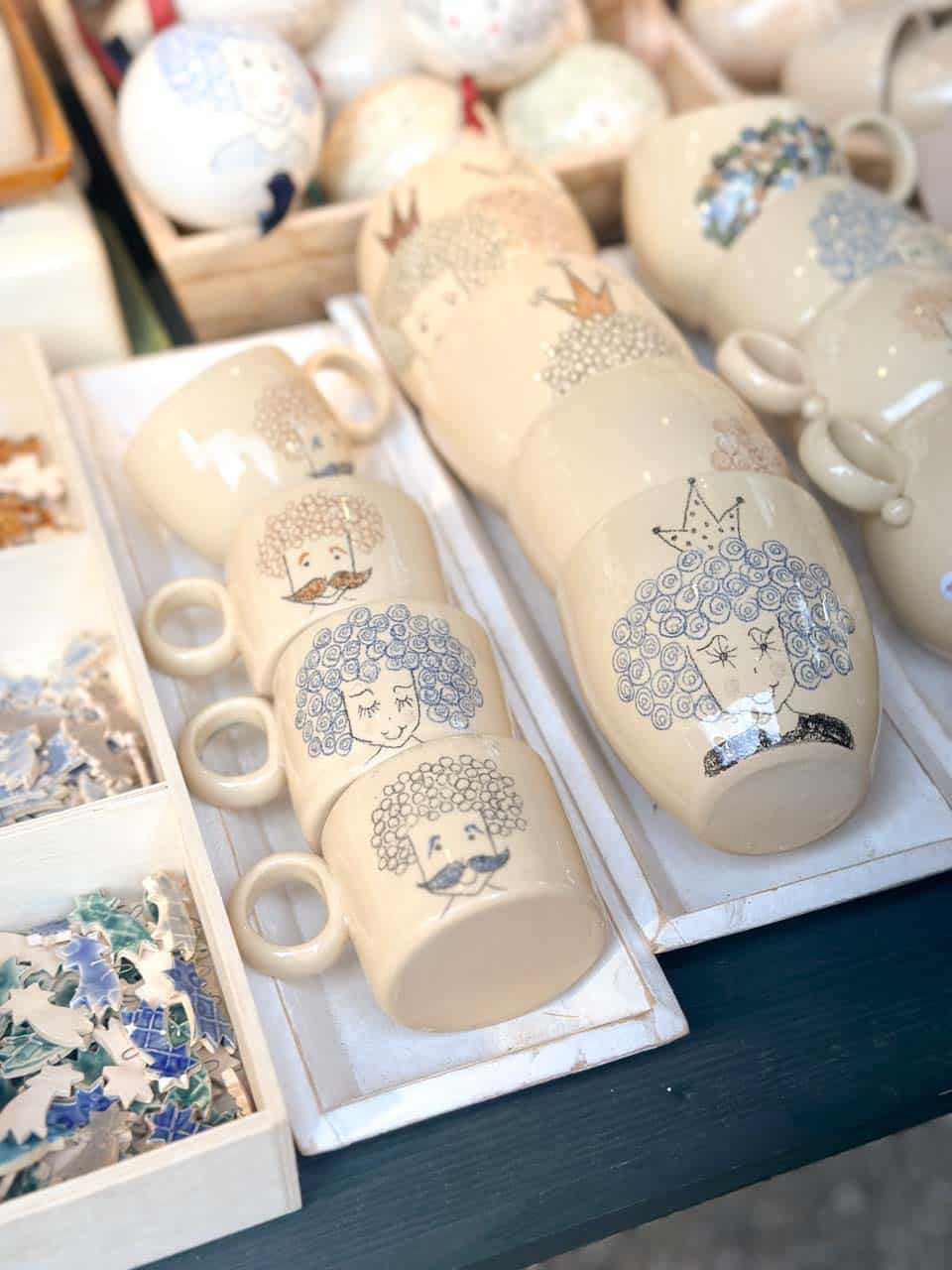 Cream-coloured ceramic mugs with hand-drawn faces and curly hair designs, displayed at a Vienna Christmas market