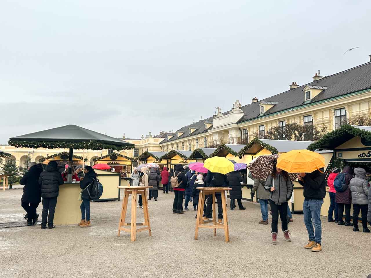 Visitors with colourful umbrellas strolling through Schönbrunn’s Christmas market on a rainy day, surrounded by decorated stalls