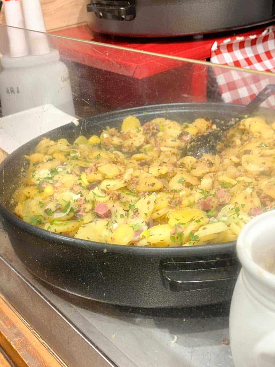 A steaming pan of golden fried potatoes with ham and herbs, displayed behind a glass counter at a food stall