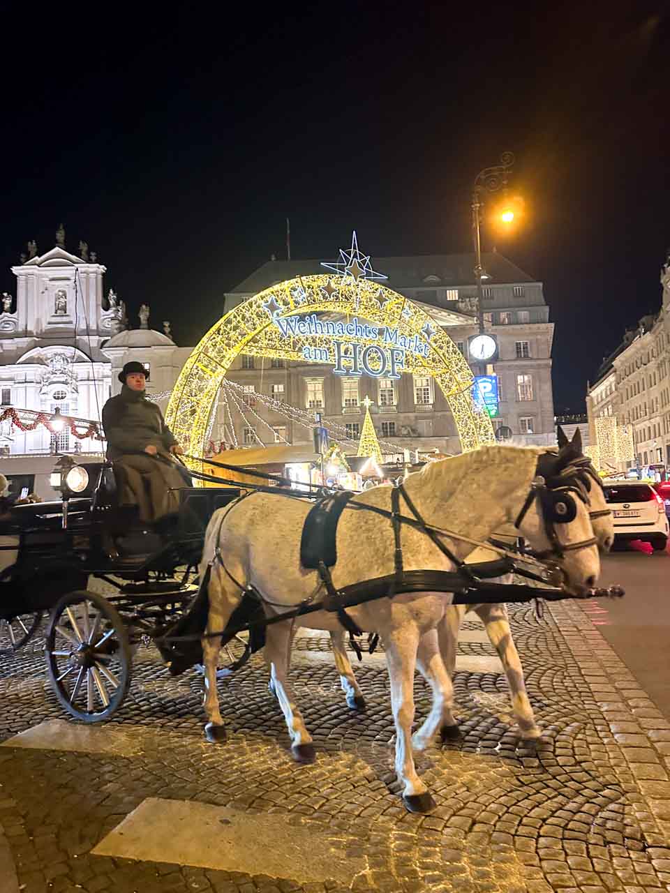 A horse-drawn carriage passing under the illuminated archway of the Weihnachts Markt am Hof at night