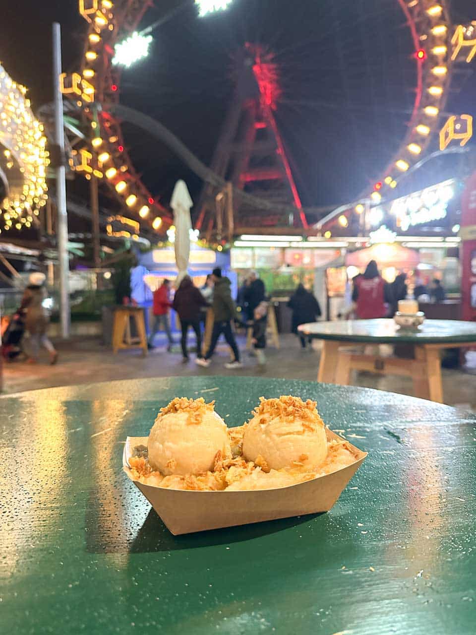 A plate of potato dumplings topped with crispy onions, with the illuminated Wiener Riesenrad Ferris wheel in the background