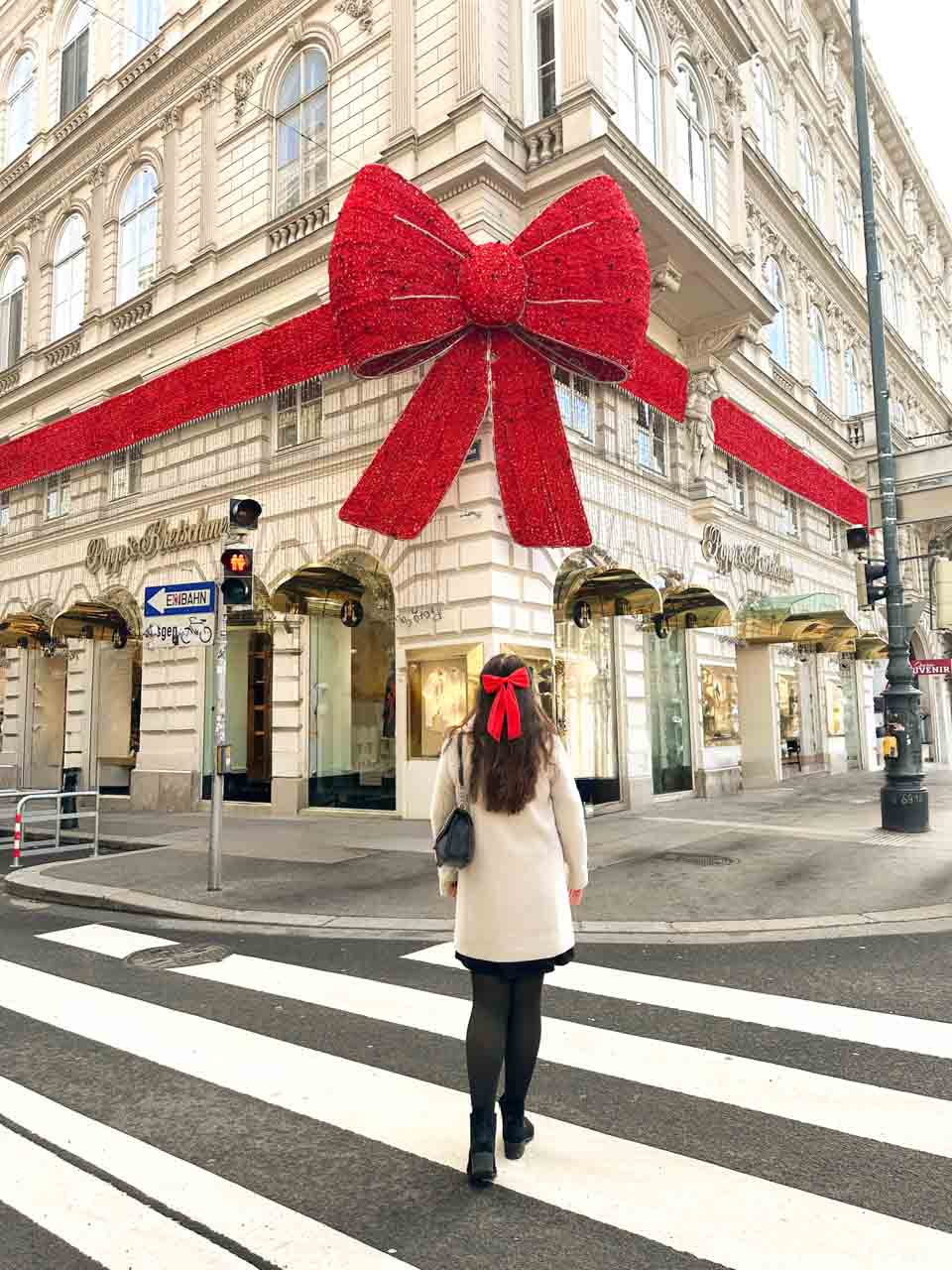 A woman in a white coat and red bow crossing the street towards the Popp & Kretschmer building in Vienna wrapped with a giant red ribbon decoration