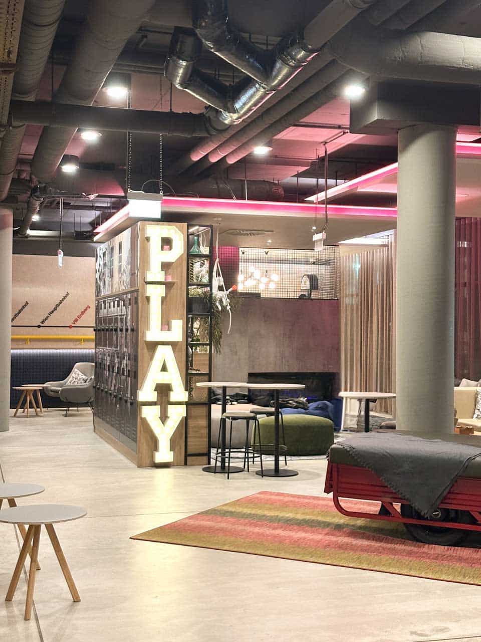 A modern lounge area with a neon “PLAY” sign, industrial decor, and cosy seating inside the Moxy Vienna City East hotel