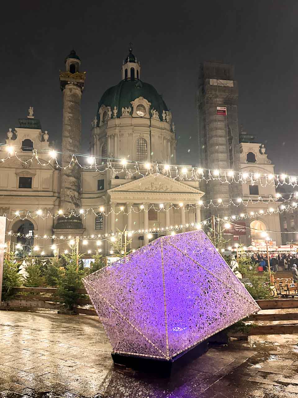 The Karlskirche illuminated at night with string lights and a glowing purple sculpture in the foreground