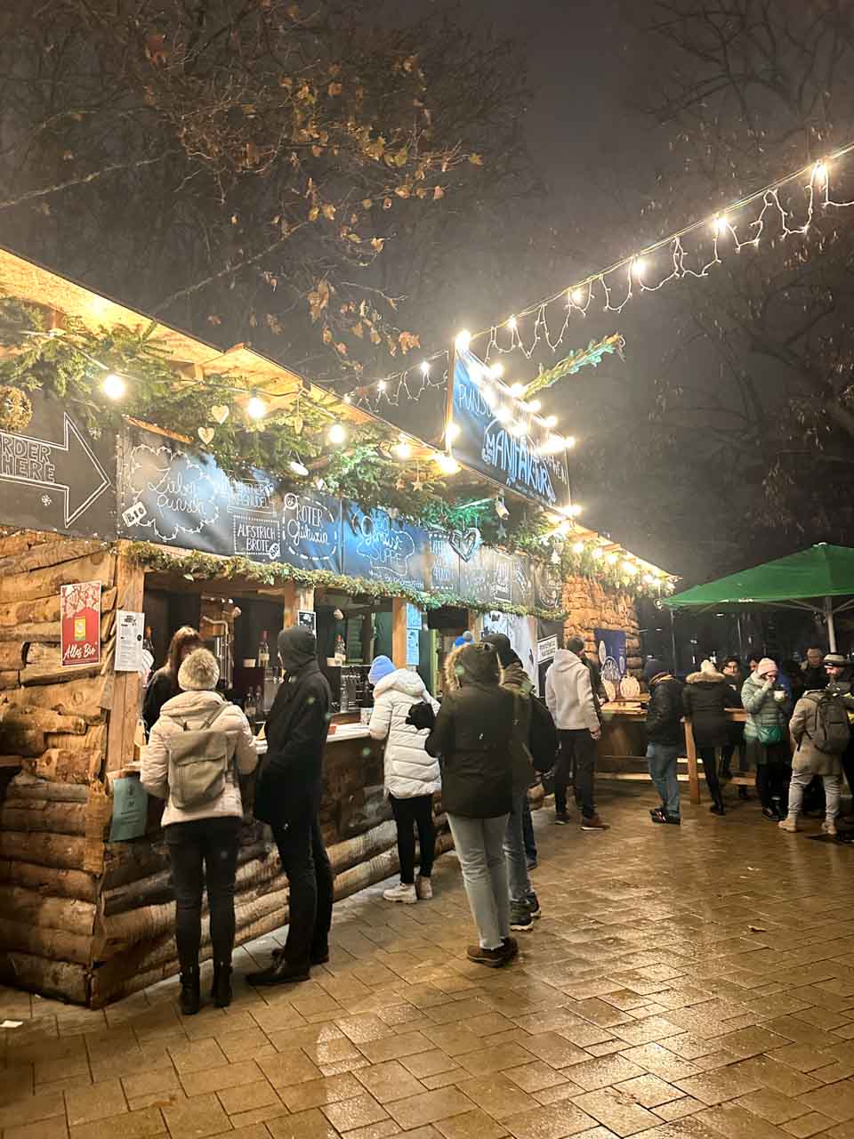 Visitors gathered around a rustic wooden food stall decorated with pine branches and string lights at the Christmas market on Karlsplatz in Vienna