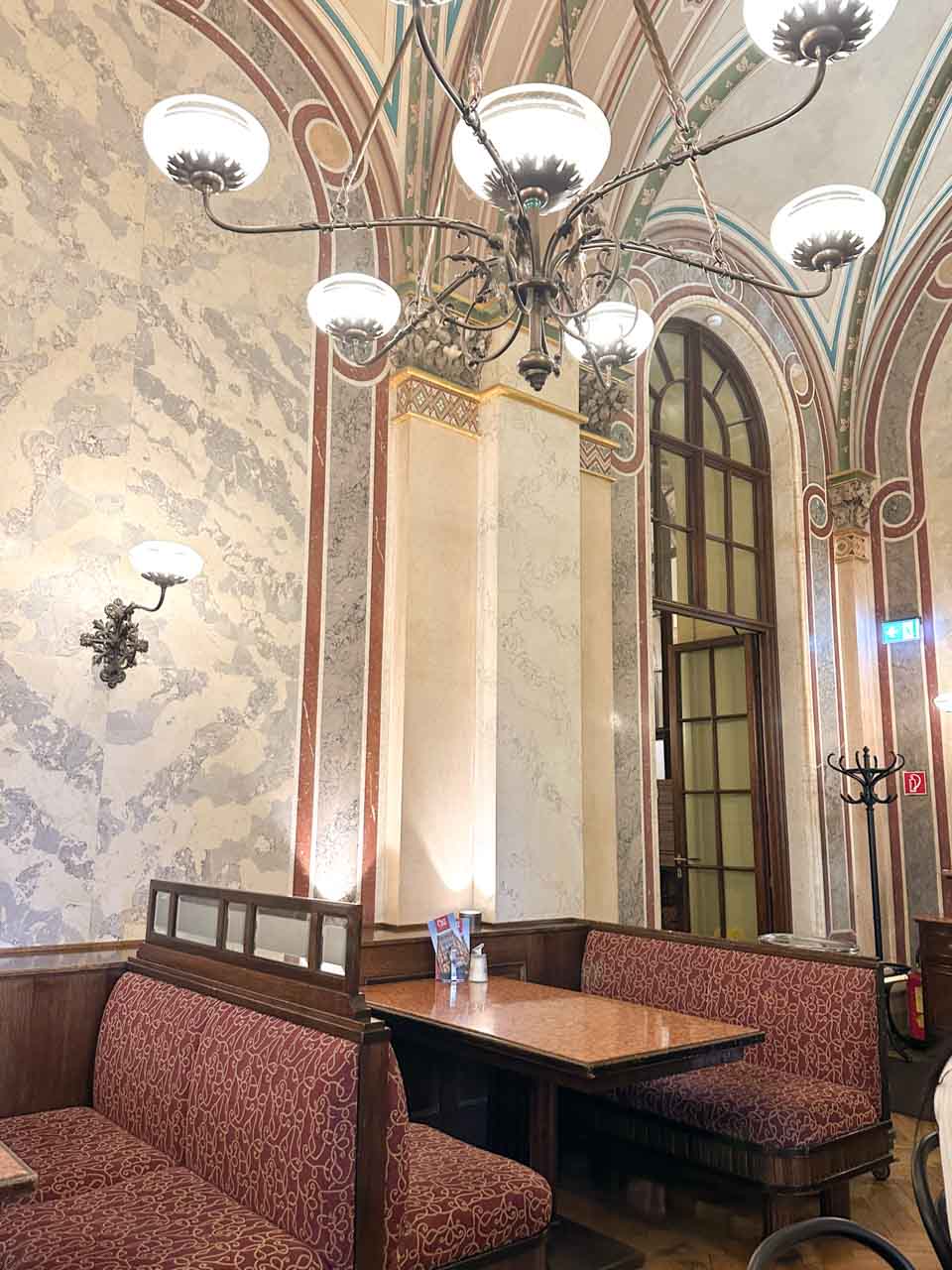 An elegant interior of Café Central in Vienna with marble walls, arched ceilings, and ornate chandeliers above cosy red upholstered booths