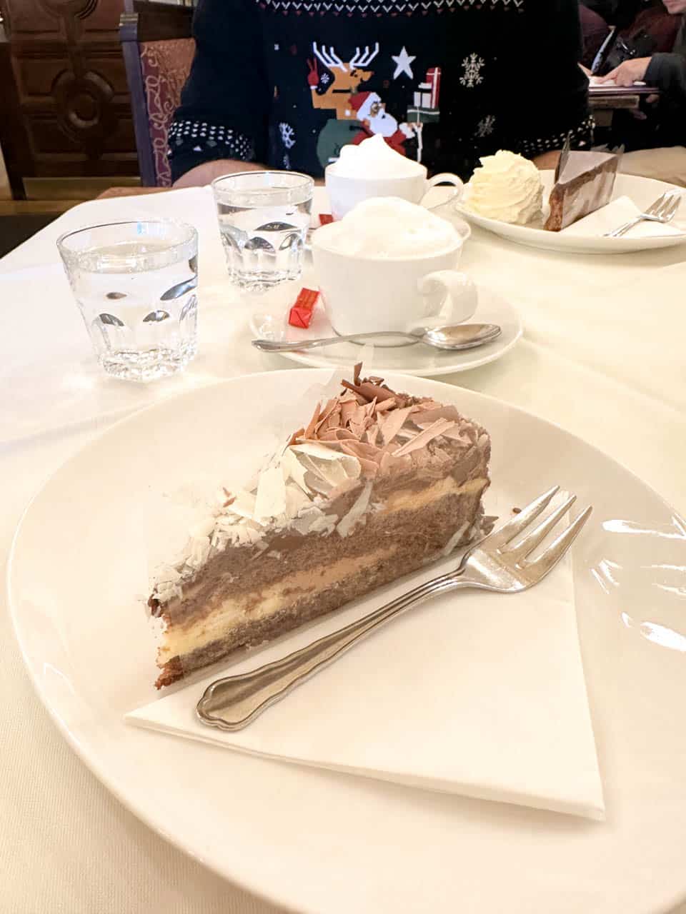 A slice of layered chocolate cake with whipped cream served on a white plate alongside a Wiener Melange coffee at Café Central