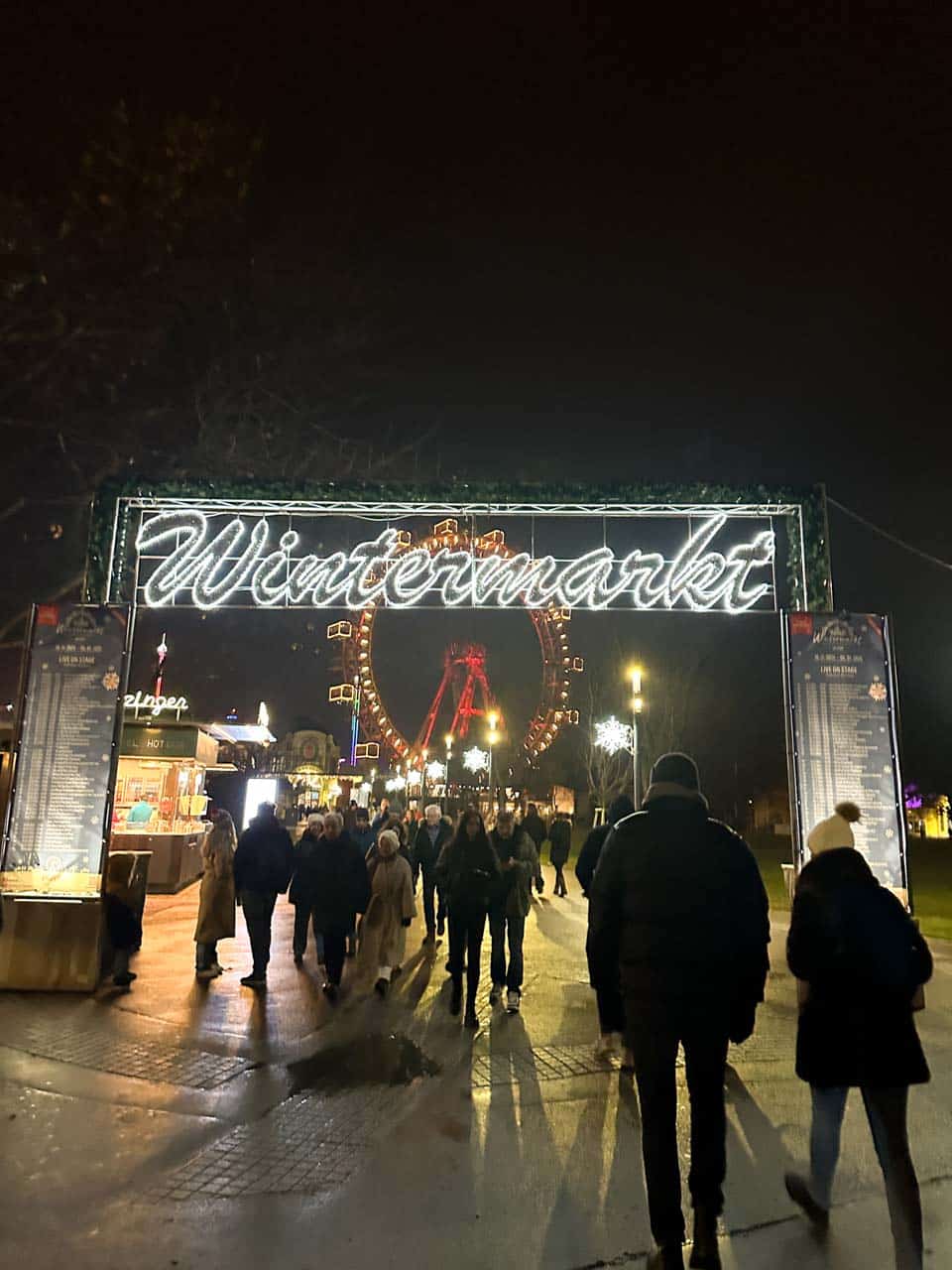 A glowing "Wintermarkt" sign at the entrance of the Prater Christmas Market in Vienna, with people walking under the festive lights