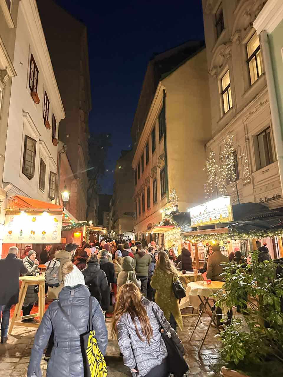 A narrow street in Vienna lined with Christmas market stalls, glowing with warm lights and filled with people enjoying the festive atmosphere
