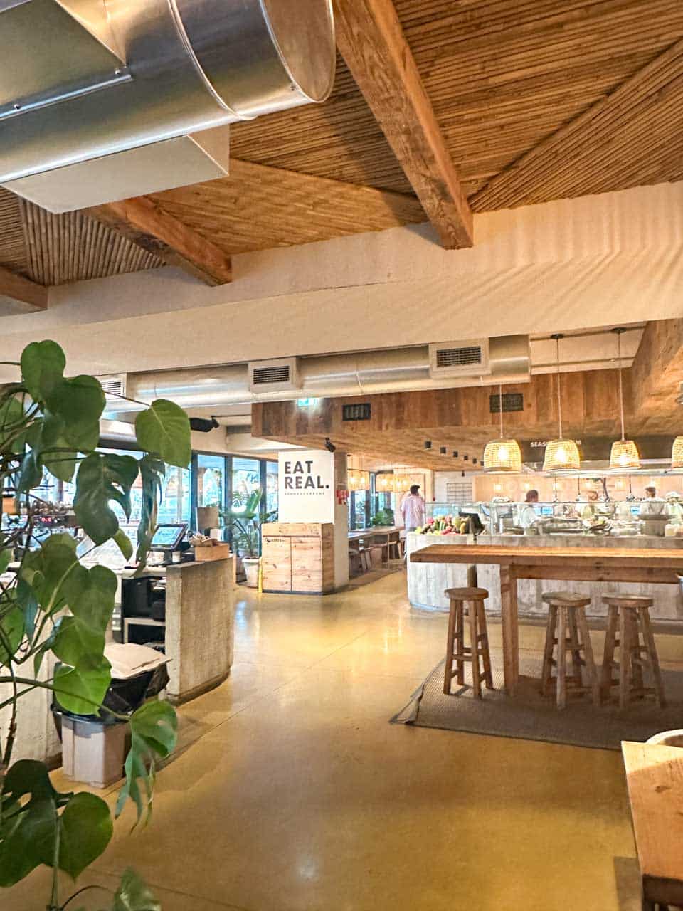The bright, airy interior of Honest Greens in Lisbon with wooden furniture, leafy plants, and a sign that says “EAT REAL” near a fresh food counter