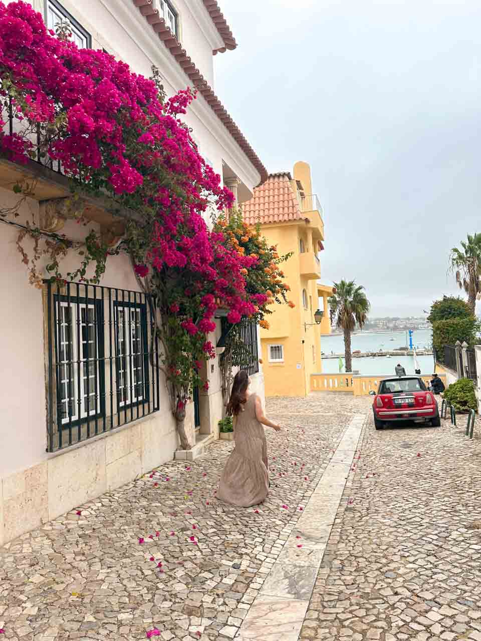 Woman in a striped dress walking down a cobbled street lined with bougainvillaea, with the ocean and palm trees in the background