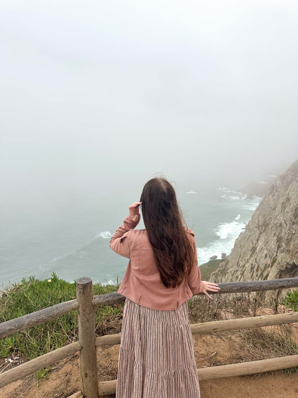 A woman standing by a wooden fence looking out at the foggy cliffs of Cabo da Roca