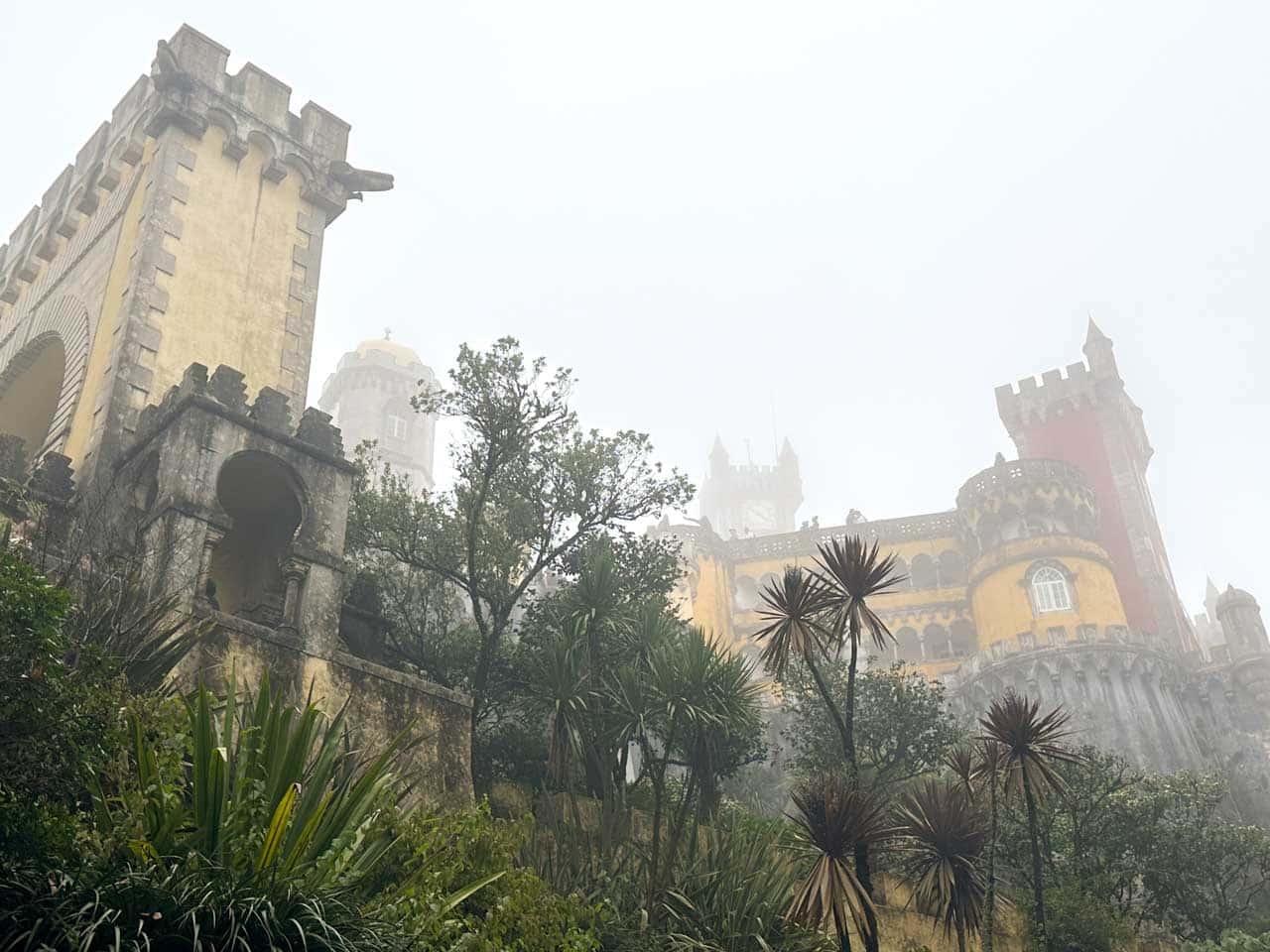 View of the misty Pena Palace in Sintra with yellow and red towers rising above palm trees and old stone arches