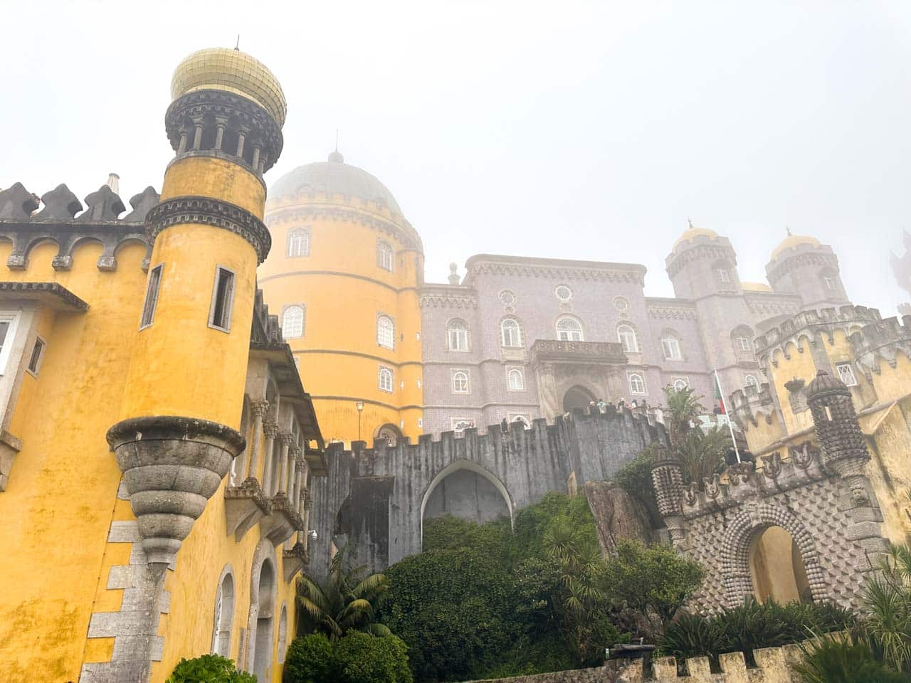 View of Pena Palace’s yellow domes and lilac walls peeking through the fog, with lush greenery below