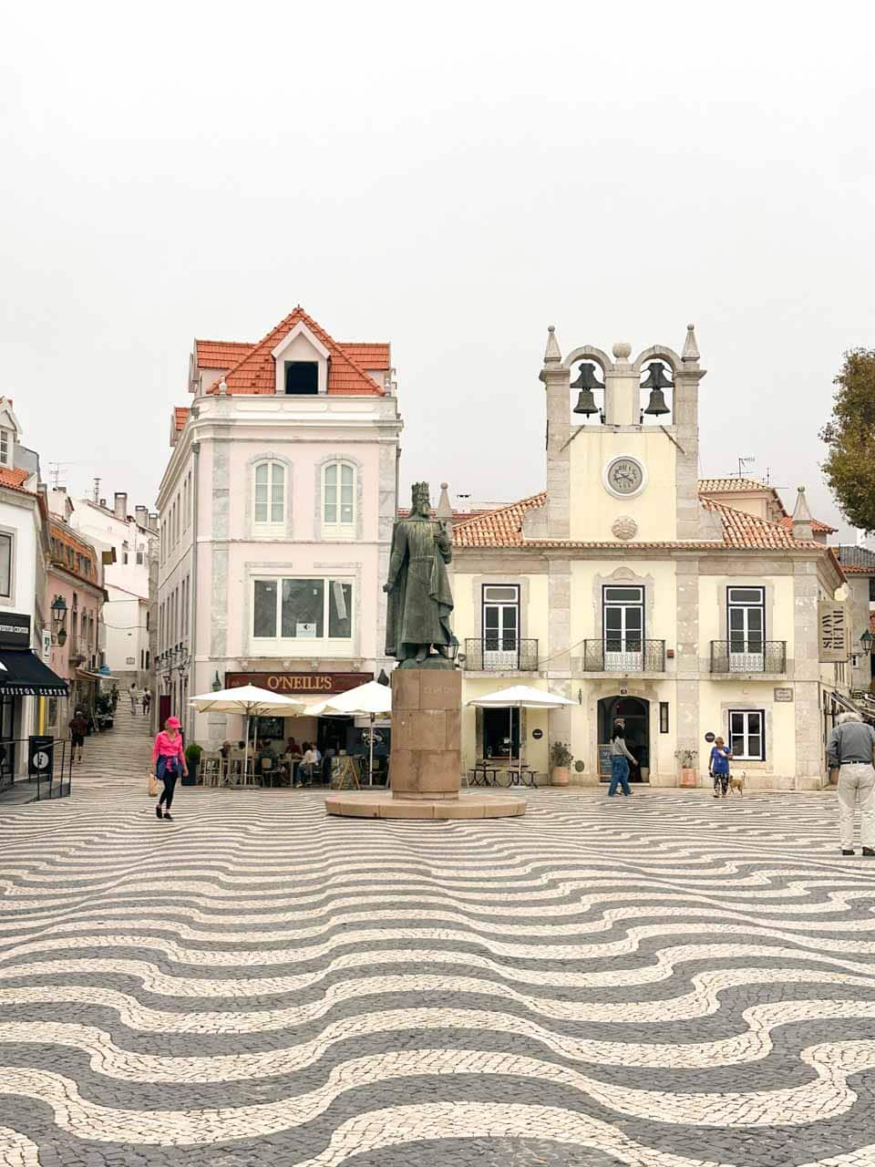 A statue standing in the centre of a square in Cascais with a wavy mosaic pavement, surrounded by cafes and whitewashed buildings