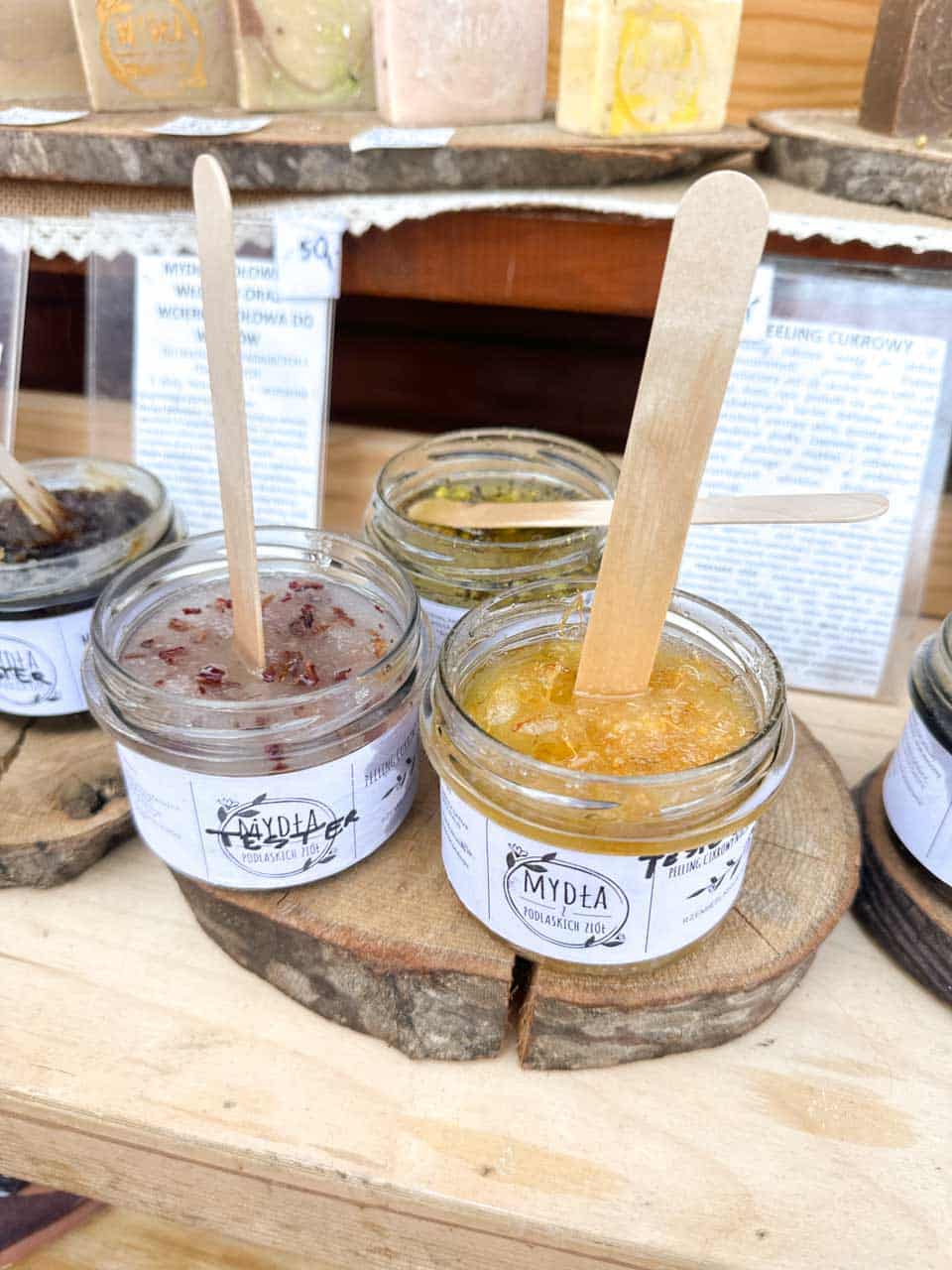 Jars of handmade body scrubs on wooden slices, each with a spatula and a label, on display at the Katowice Easter Market
