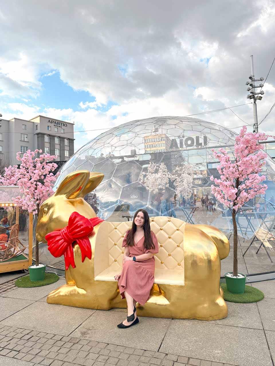 A woman in a pink dress sitting on a golden bunny bench, with cherry blossom trees and a dome behind her