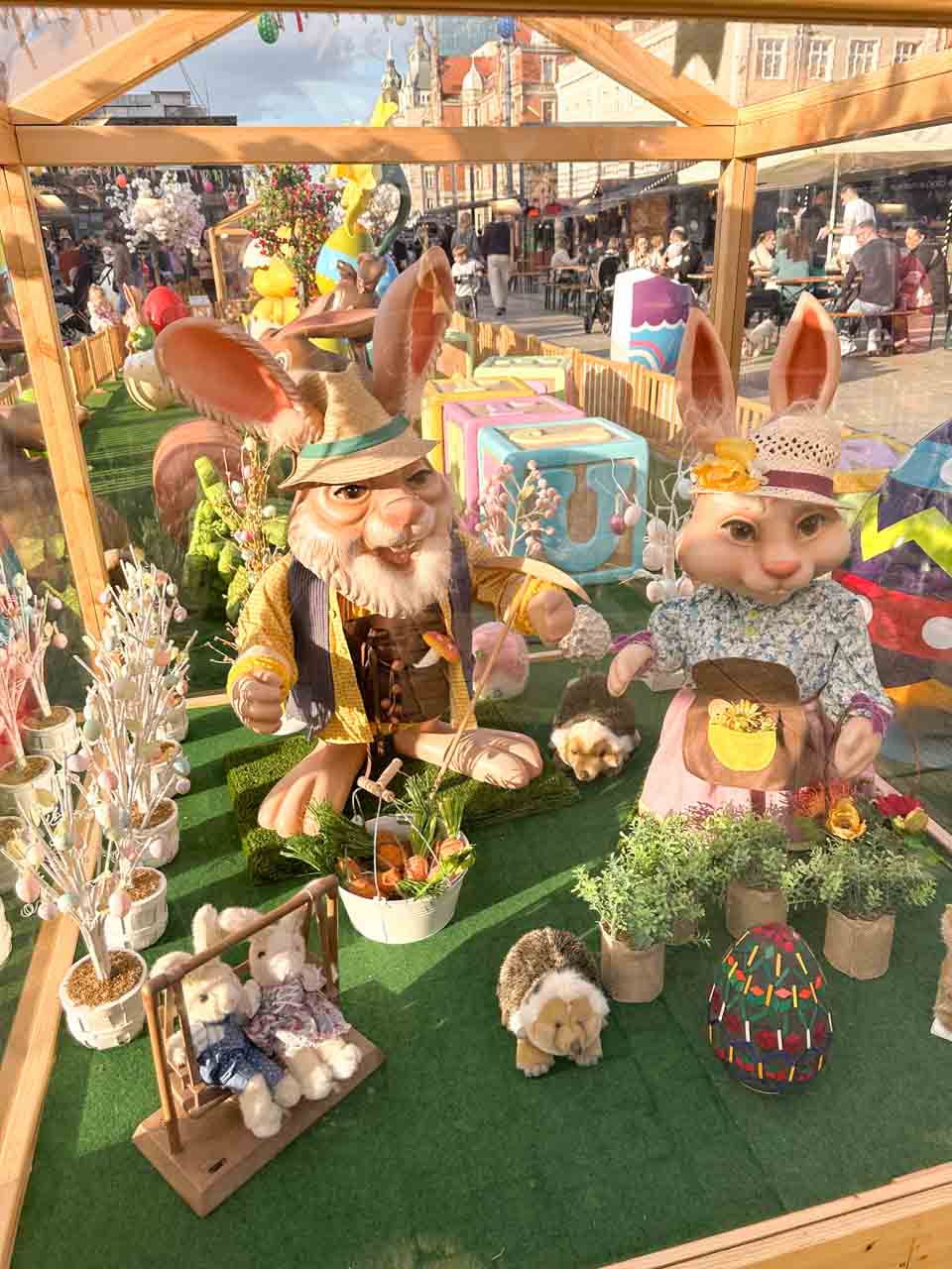 Large Easter bunny figures dressed in clothes, surrounded by flowers, eggs, and cute animal toys in a glass display in the Market Square in Katowice