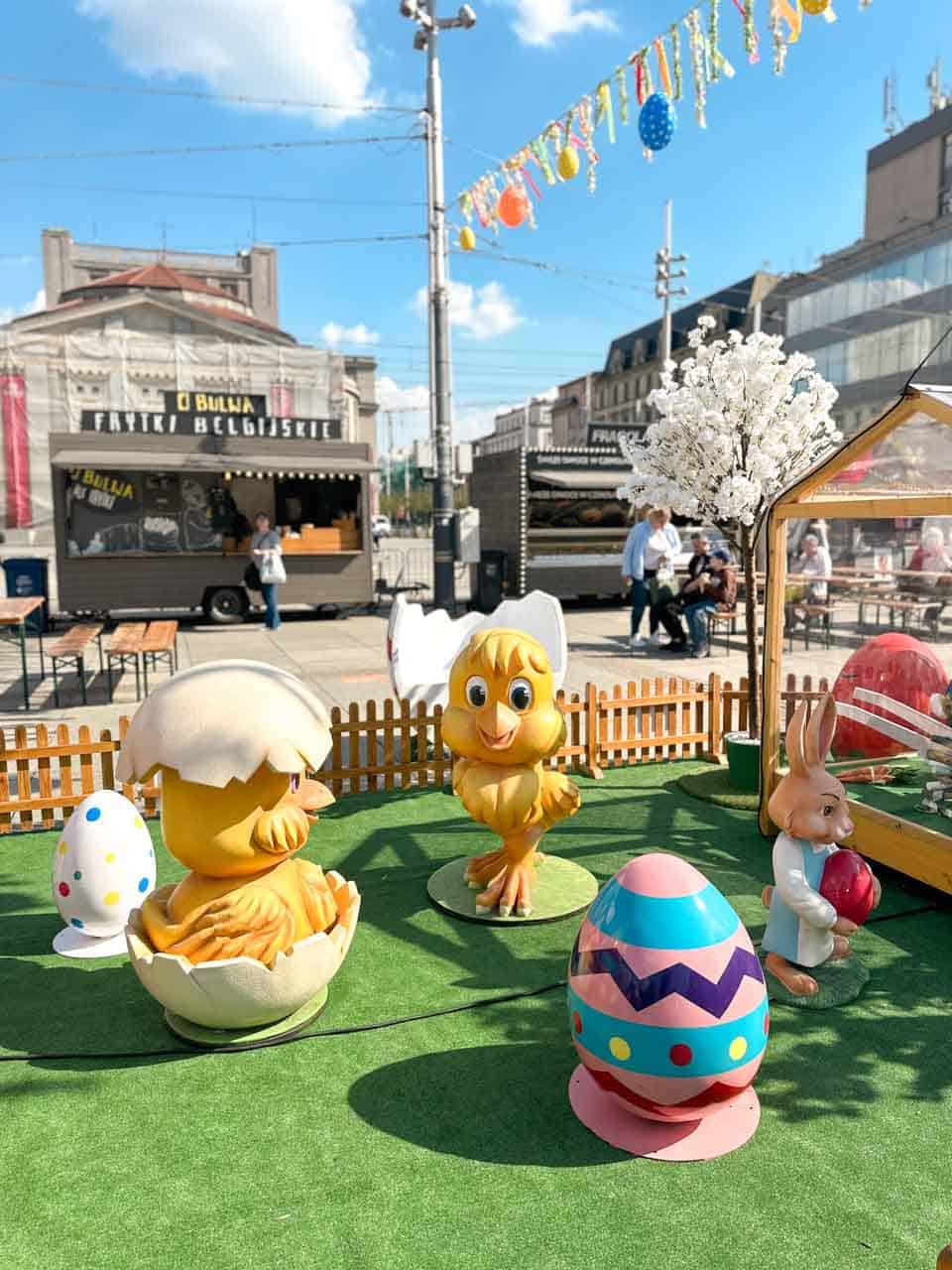 Large chick and bunny figures surrounded by painted eggs, with food stalls and Easter decorations behind