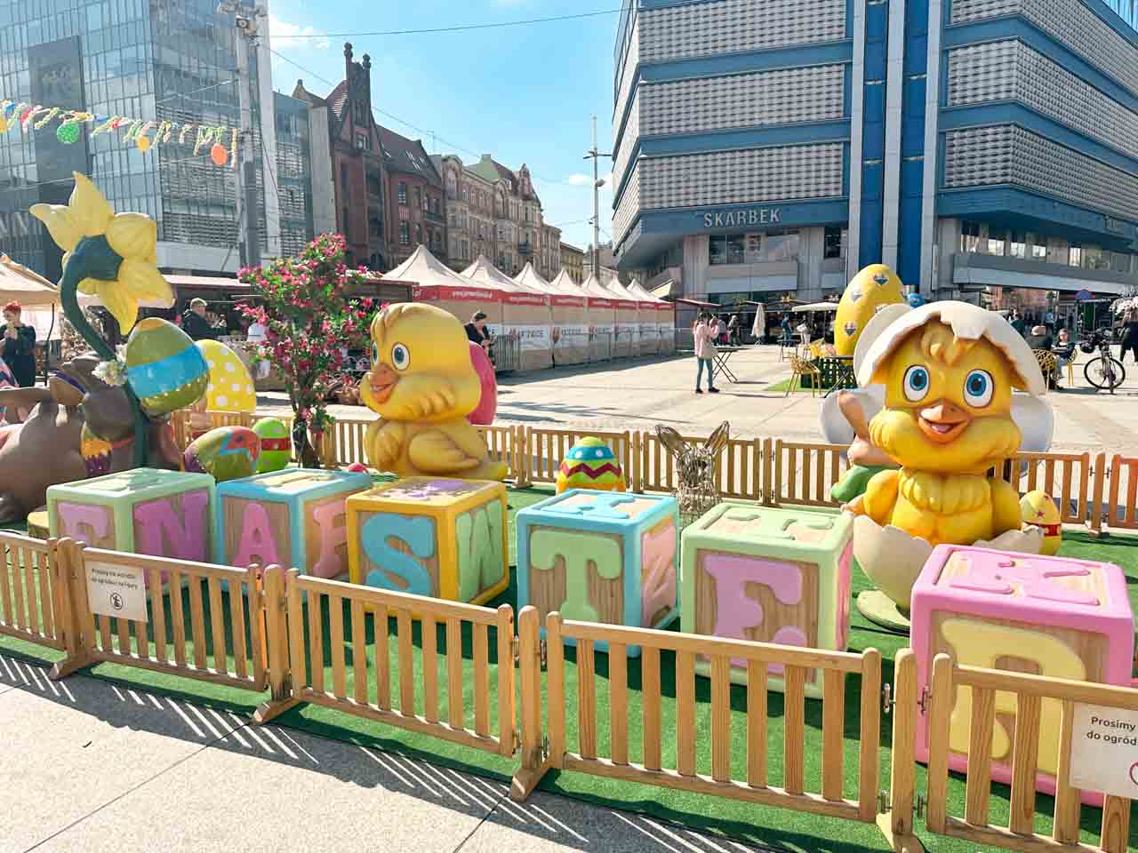 Big yellow chicks and Easter eggs displayed behind pastel toy blocks, set up in a fenced area in the Market Square in Katowice, Poland