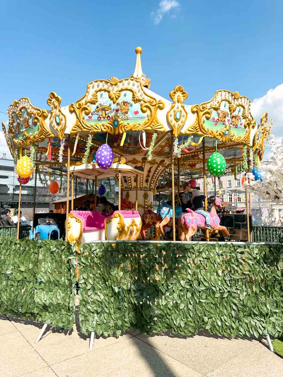 A colourful carousel decorated with Easter eggs and ribbons, with kids riding painted horses under a clear blue sky
