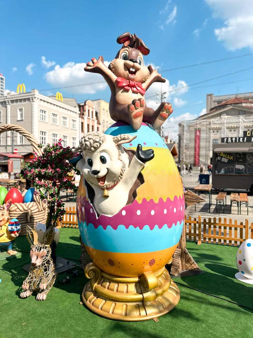 A cheerful Easter display with a bunny and lamb bursting out of a giant painted egg, surrounded by flowers and decorations