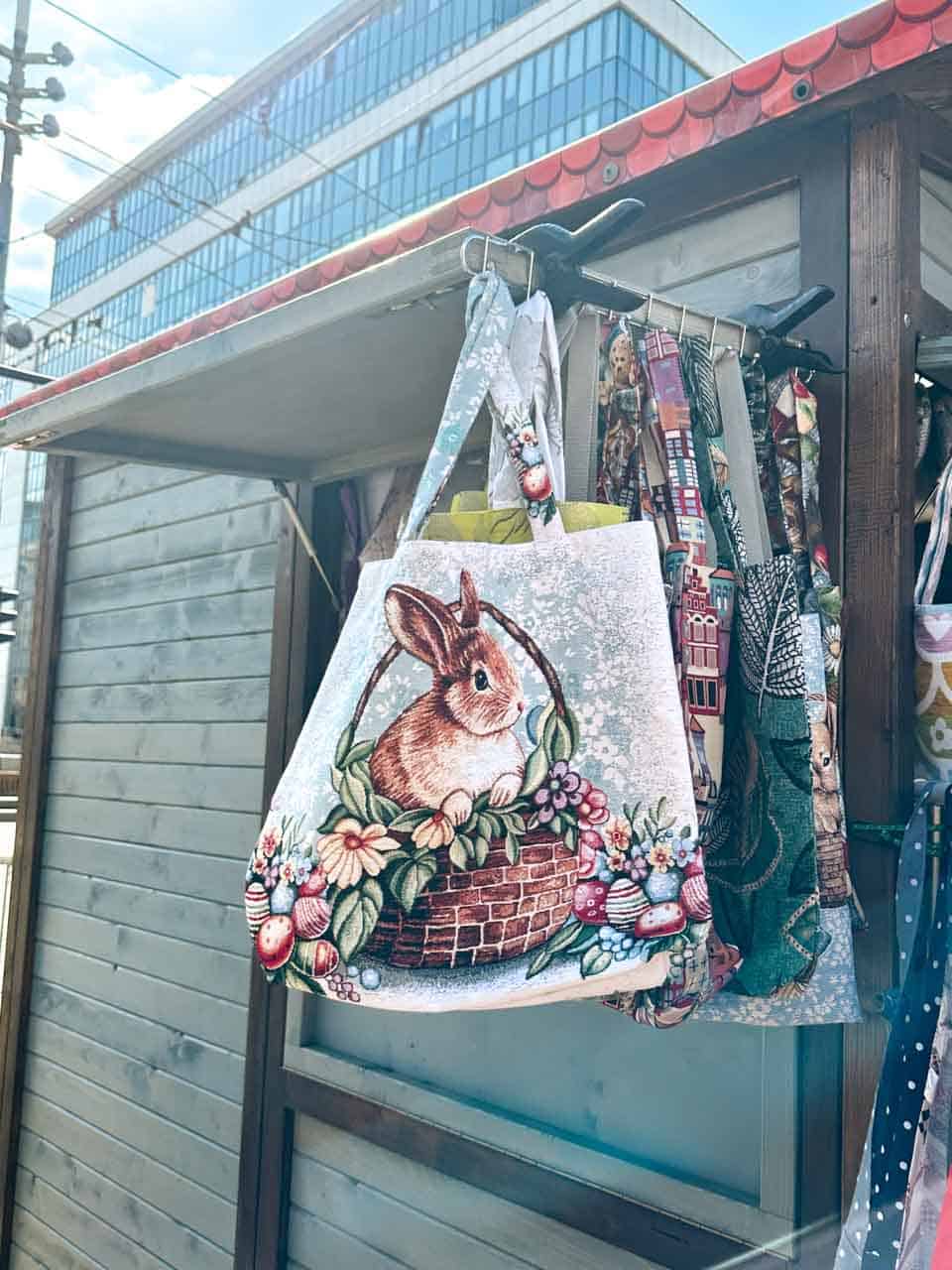 A fabric tote bag with a cute Easter bunny in a basket, decorated with painted eggs and flowers, hanging on a wooden stall in Katowice, Poland