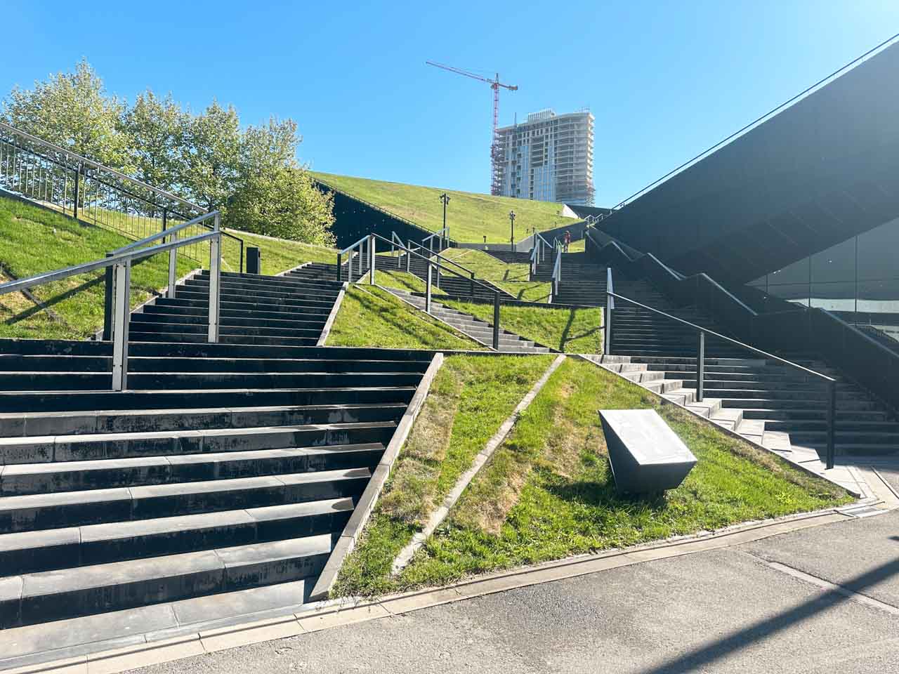 Modern outdoor stairs leading up a grassy hill in Katowice, with a crane and a tall building in the background