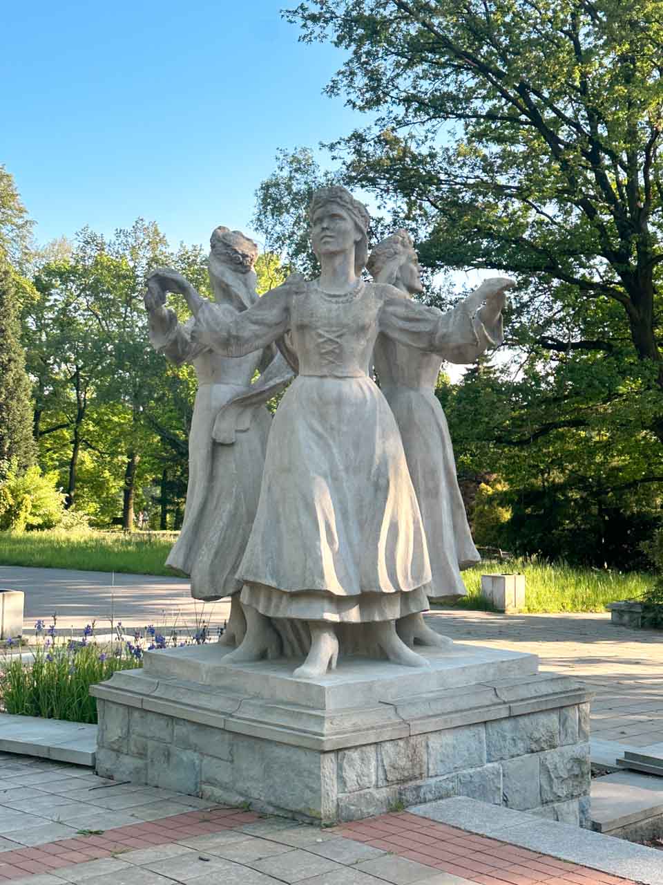 Stone statue of three women in traditional dress dancing in a circle, set inside Silesia Park