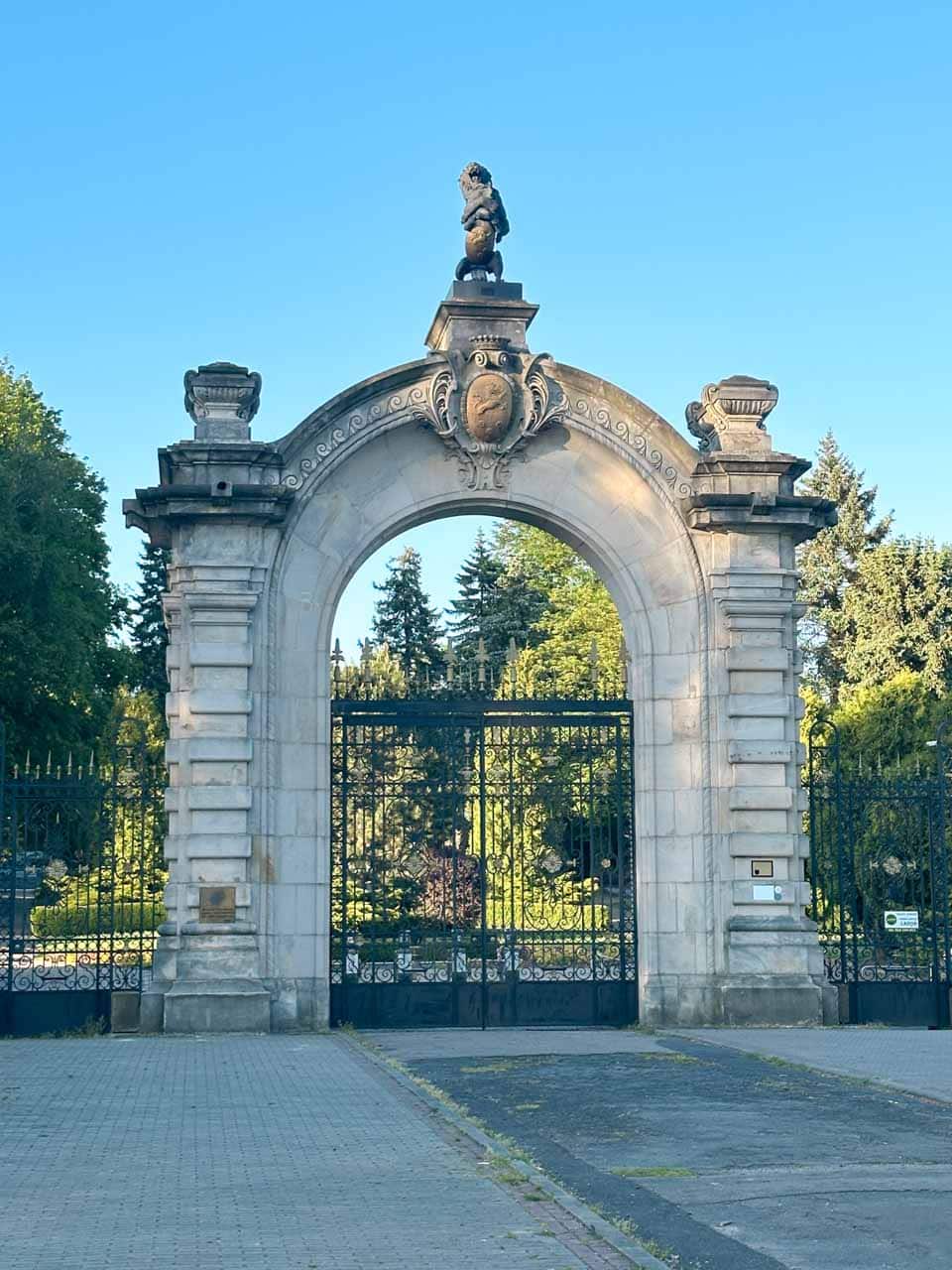 A grand stone gate with a lion statue on top, leading to the Silesian Zoo with trees and greenery in the background