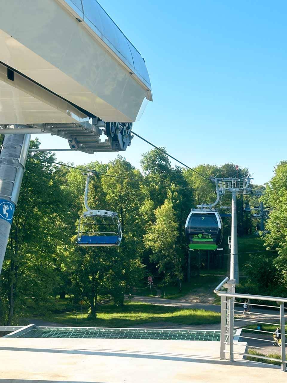 Cable cars and chairlifts travelling over a green park area, with trees and a bike path below