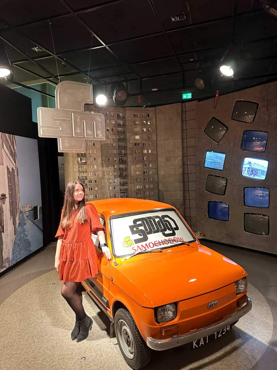 A woman in an orange dress standing next to an orange vintage Fiat 126p inside the Silesian Museum in Katowice, Poland