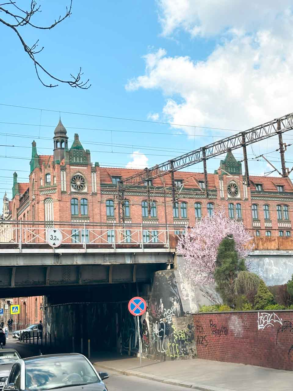 A red-brick building with green rooftops seen behind a railway bridge covered in graffiti and spring blossoms in Katowice, Poland